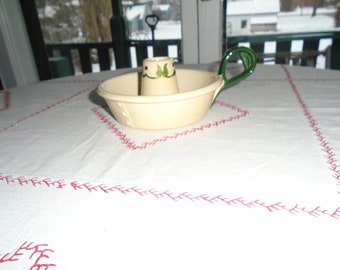 May include: A white ceramic candle holder with a green handle and green floral design. The holder is sitting on a white tablecloth with pink stitching.