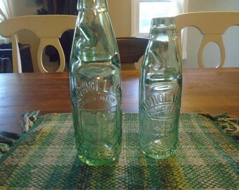 May include: Two green glass bottles with a textured surface and the words "Nichol's Lager Trade Mark" embossed on the front. The bottles are sitting on a green and white checkered placemat.