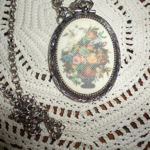 May include: A silver chain necklace with an oval pendant featuring a floral embroidery design. The pendant has a silver frame and a white background.