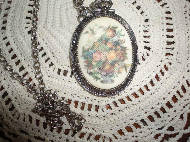 May include: A silver chain necklace with an oval pendant featuring a floral embroidery design. The pendant has a silver frame and a white background.