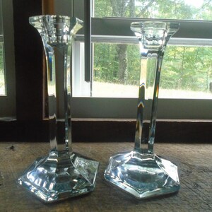 May include: Two clear glass candle holders with a square base and a tall, slender design. The candle holders are made of clear glass and have a polished finish.