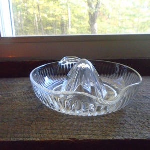 May include: A clear glass citrus juicer with a ribbed design. The juicer has a central cone with three sections for squeezing citrus fruits.