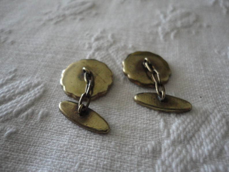 May include: A pair of gold cufflinks with a chain connecting them. The cufflinks have a round design with a textured surface.