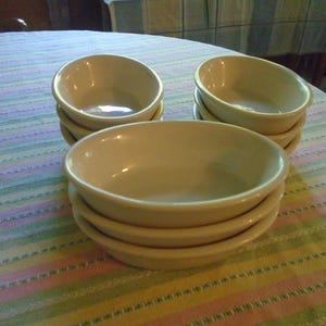 May include: A stack of six off-white ceramic bowls, oval-shaped with a slight lip. Three stacks of bowls are arranged on a striped tablecloth with pastel colors. The bowls are empty and appear clean.
