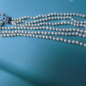 May include: A multi-strand pearl necklace with a silver clasp. The necklace is made of white pearls and is laid out on a turquoise surface.