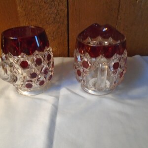 May include: A set of two clear glass sugar and creamer with a ruby red overlay. The glass has a diamond pattern and is accented with small red dots.