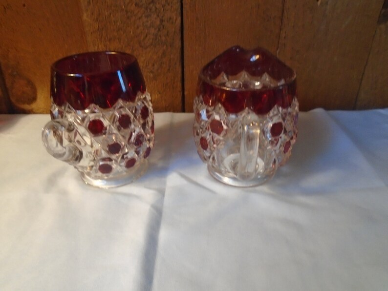 May include: A set of two clear glass sugar and creamer with a ruby red overlay. The glass has a diamond pattern and is accented with small red dots.