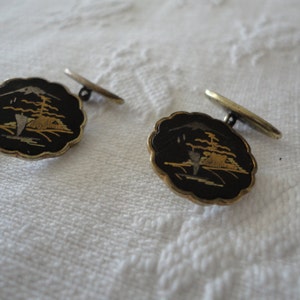 May include: A pair of cufflinks with a black background and gold and silver accents. The cufflinks feature a stylized design of a mountain scene.