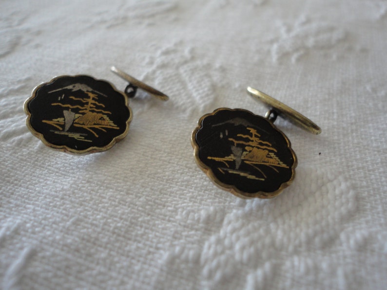 May include: A pair of cufflinks with a black background and gold and silver accents. The cufflinks feature a stylized design of a mountain scene.