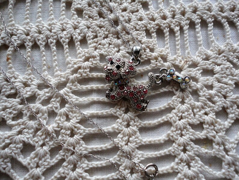 Pu&ograve; includere: Una collana con catena d'argento con un ciondolo a forma di cane in strass rosa e un ciondolo a forma di osso in strass abbinato.