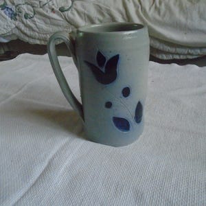 May include: A tall, light blue ceramic mug with a curved handle. The mug features a dark blue floral design with a tulip and leaf pattern. The mug is sitting on a white, textured surface.