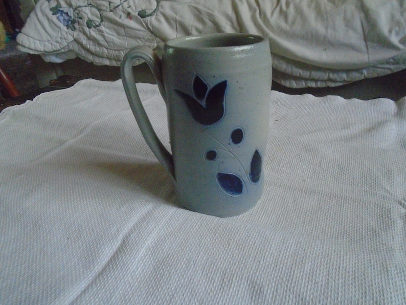 May include: A tall, light blue ceramic mug with a curved handle. The mug features a dark blue floral design with a tulip and leaf pattern. The mug is sitting on a white, textured surface.