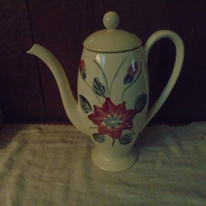 May include: Cream-colored ceramic teapot with a floral design. The teapot features a large red flower with blue and green leaves. It has a curved spout, a handle, and a lid with a small knob. The teapot is likely vintage.