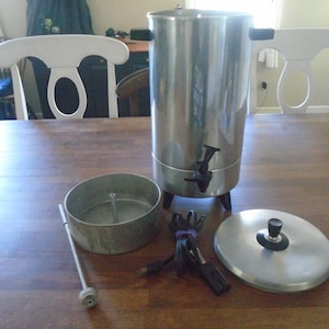 May include: A silver coffee maker with a black handle and a black cord. The coffee maker is disassembled and laid out on a wooden table. The parts include a filter basket, a lid, and a cord.