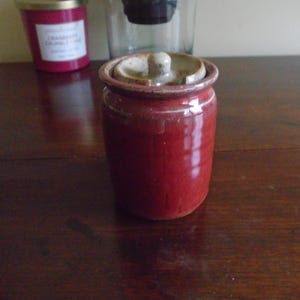 May include: A small, red ceramic jar with a lid. The jar has a cylindrical shape and a slightly rough texture. The lid is a lighter shade of brown and has a small knob on top. The jar is sitting on a dark wooden surface.