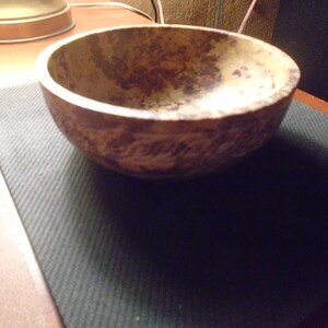 May include: A rustic, handcrafted bowl with a textured, mottled brown and tan finish. The bowl has a rounded shape and sits on a dark green surface. The interior of the bowl is a lighter tan color.