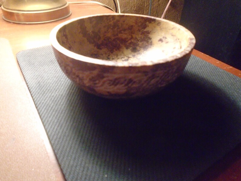 May include: A rustic, handcrafted bowl with a textured, mottled brown and tan finish. The bowl has a rounded shape and sits on a dark green surface. The interior of the bowl is a lighter tan color.