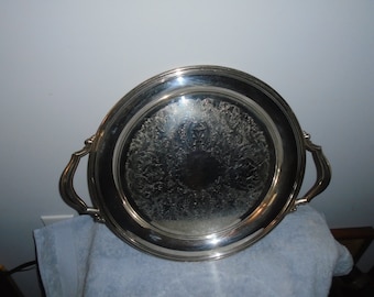 May include: A silver-colored serving tray with ornate floral detailing in the center. The tray has two curved handles and a raised rim. The tray is round and appears to be made of metal.
