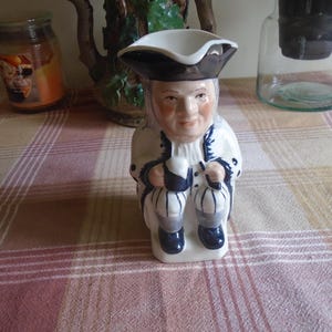 May include: A ceramic figurine of a man in a tricorn hat and colonial attire. The figurine is predominantly white with blue and black accents. It is holding a lantern and a mug. The figurine is sitting on a checkered tablecloth.