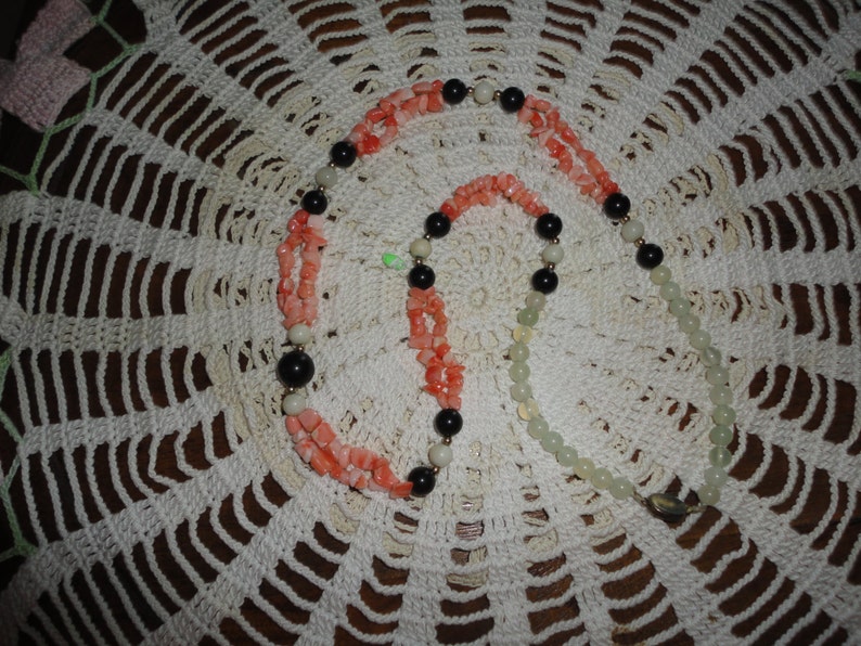 Puede incluir: Un collar de varias hebras con cuentas de coral, blancas y negras. El collar est&aacute; sobre un encaje de ganchillo blanco.