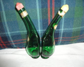 May include: Two green glass bottles with unique shapes and colorful stoppers. The bottles are empty and have a vintage look.