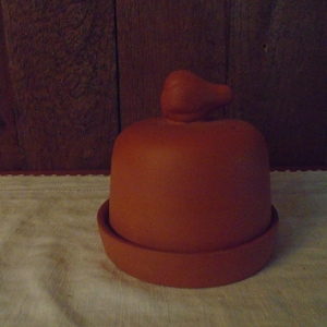 May include: A terracotta-colored butter dish with a lid and a small handle on top. The dish has a round base and a domed lid. The handle is shaped like a small bird. The butter dish is sitting on a white surface.