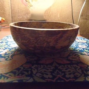May include: A textured, brown wooden bowl with a wide rim. The bowl sits on a blue patterned surface with floral and bird designs. The background includes a lamp and wooden paneling.