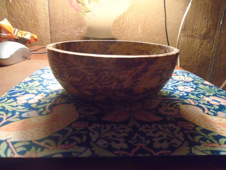 May include: A textured, brown wooden bowl with a wide rim. The bowl sits on a blue patterned surface with floral and bird designs. The background includes a lamp and wooden paneling.