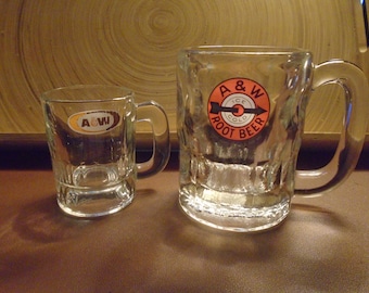 May include: Two glass A&W Root Beer mugs. The larger mug has a handle and the smaller mug does not. Both mugs have the A&W logo with the text "Ice Cold Root Beer".