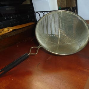 May include: A stainless steel mesh strainer with a black handle. The strainer has a round shape and a fine mesh. The handle is attached to the strainer with a metal bracket. The strainer is on a wooden surface.