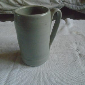 May include: A tall, light gray ceramic mug with a curved handle. The mug has a cylindrical shape with a slightly flared top and a simple, minimalist design. The mug is sitting on a white, textured surface.