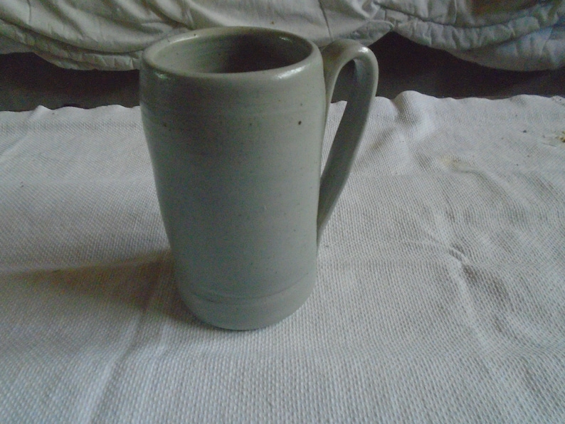 May include: A tall, light gray ceramic mug with a curved handle. The mug has a cylindrical shape with a slightly flared top and a simple, minimalist design. The mug is sitting on a white, textured surface.