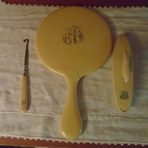 May include: An antique vanity set in a cream color, including a round hand mirror, a small brush, and a crochet hook. Each piece has a matching monogram in a dark color. The set is displayed on a light-colored fabric.