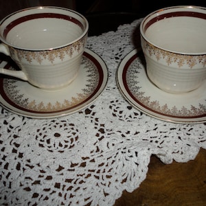 May include: Two white teacups with gold and red trim and matching saucers on a white crocheted doily.