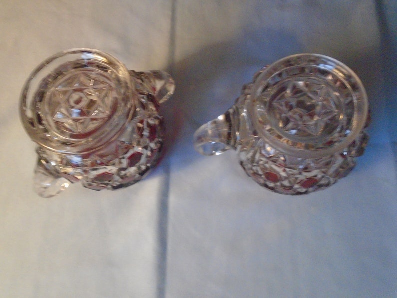 May include: Two clear glass cream jugs with a red, star-shaped pattern. The jugs have a round, clear glass top with a star-shaped design.