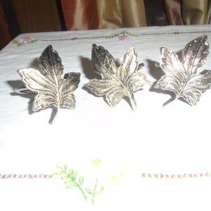 May include: Three silver-toned metal napkin rings, each featuring a detailed, embossed maple leaf design. The leaves have defined veins and serrated edges, with a metallic finish that reflects light, creating highlights and shadows across the surface.