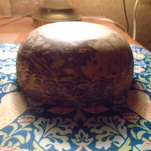 May include: A round, brown decorative bowl with a carved floral design around the base. The bowl is set on a blue and white patterned surface with birds and floral motifs. The bowl has a light-colored top.