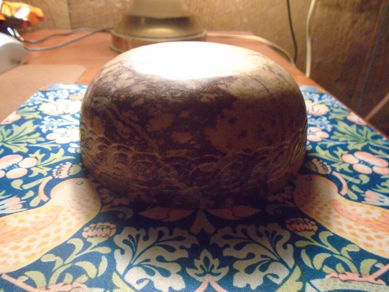 May include: A round, brown decorative bowl with a carved floral design around the base. The bowl is set on a blue and white patterned surface with birds and floral motifs. The bowl has a light-colored top.