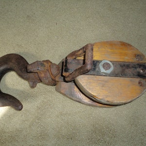 May include: A vintage wooden pulley with a metal hook and a black metal band. The pulley is made of two pieces of wood that are joined together with metal screws.
