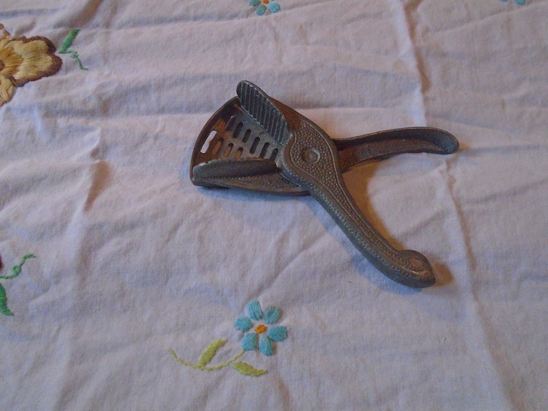 May include: A silver metal citrus juicer with a handle and a gridded surface. The juicer is lying on a white tablecloth with embroidered flowers.