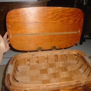 Puede incluir: Una cesta de picnic de madera vintage con tapa con bisagras e interior de cesta tejido. La tapa es de madera y tiene un ribete tejido.