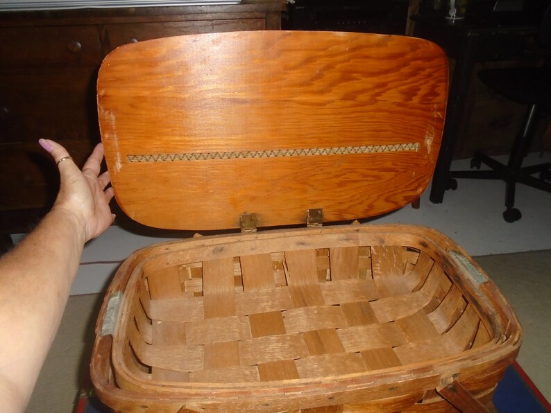 Puede incluir: Una cesta de picnic de madera vintage con tapa con bisagras e interior de cesta tejido. La tapa es de madera y tiene un ribete tejido.