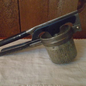 May include: An antique potato ricer with a black handle and a perforated metal bowl. The ricer is made of metal and has a lever-style design. The handle is black, and the bowl is silver. The ricer is designed for mashing potatoes.