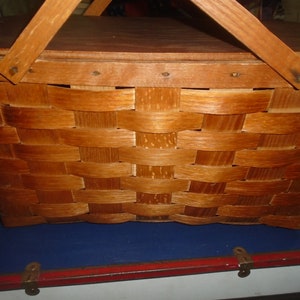 Puede incluir: Una cesta de picnic de madera vintage con un dise&ntilde;o tejido y una tapa con bisagras. La cesta tiene un tinte marr&oacute;n y dos asas de madera.