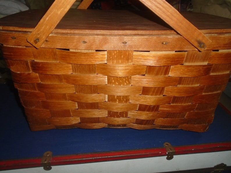 Puede incluir: Una cesta de picnic de madera vintage con un dise&ntilde;o tejido y una tapa con bisagras. La cesta tiene un tinte marr&oacute;n y dos asas de madera.