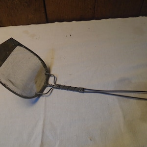 May include: A vintage metal kitchen utensil with a wire handle and a flat, round mesh head. The mesh head is made of a woven material and has a dark gray color.