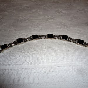 May include: A silver bracelet with black square stones. The bracelet has a clasp closure.