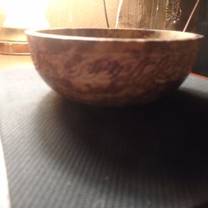 May include: A brown, round bowl with a textured surface and a decorative pattern. The bowl is made of a light-colored material and sits on a dark surface. The bowl is likely used for serving food or as a decorative item.