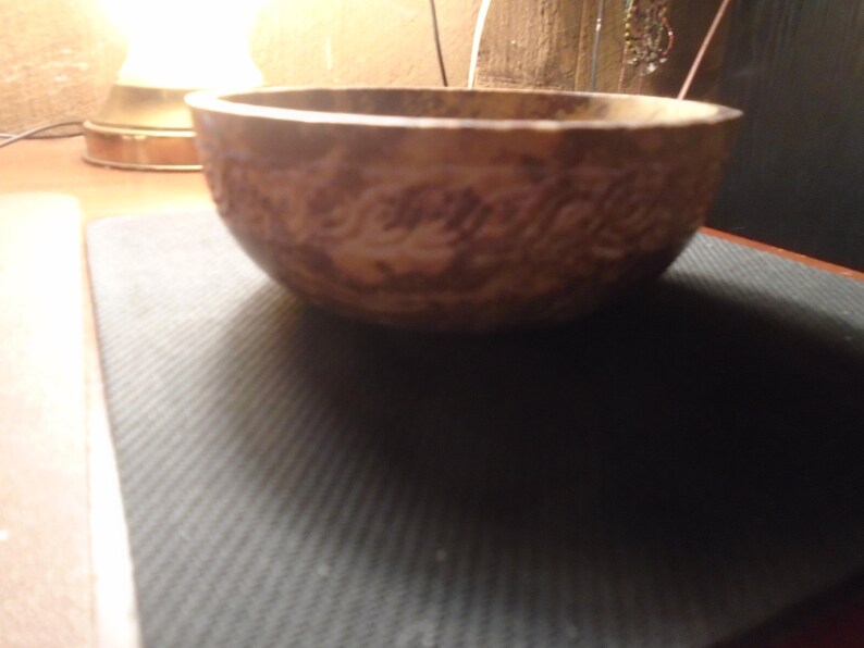 May include: A brown, round bowl with a textured surface and a decorative pattern. The bowl is made of a light-colored material and sits on a dark surface. The bowl is likely used for serving food or as a decorative item.