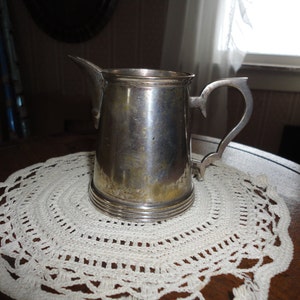 May include: A silver creamer with a rounded body and a handle. The creamer is sitting on a white crocheted doily.